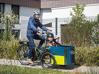 Un homme à vélo portant un casque transporte à l'avant du vélocargo estampillé Vélila 2 enfants. Ils sont en sécurité dans un