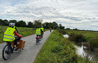 Plusieurs personnes se baladent à vélo sur une piste cycclable avec gilet fluo de sécurité et casqu. UN des vélo est un vélo cargo qui permet de transporter deux enfants à l'avant du vélo en toute sécurité. - Agrandir l'image, .JPG 2 Mo (fenêtre modale)